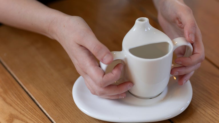 A close-up image of a person holding an accessible, ergonomic white cup with two hands on a wooden table. The cup features a spout and is placed on a matching saucer, designed to assist individuals with limited mobility or special needs. The calm, indoor setting of a café can be inferred from the soft lighting and simple wooden surface.